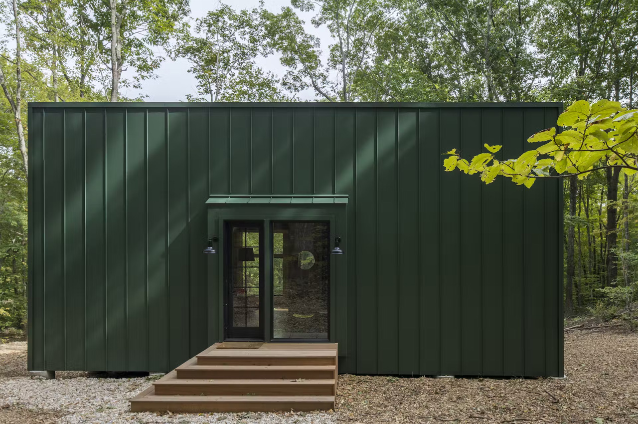 This dark green cabin floats above a sloping terrain in a forest in Connecticut