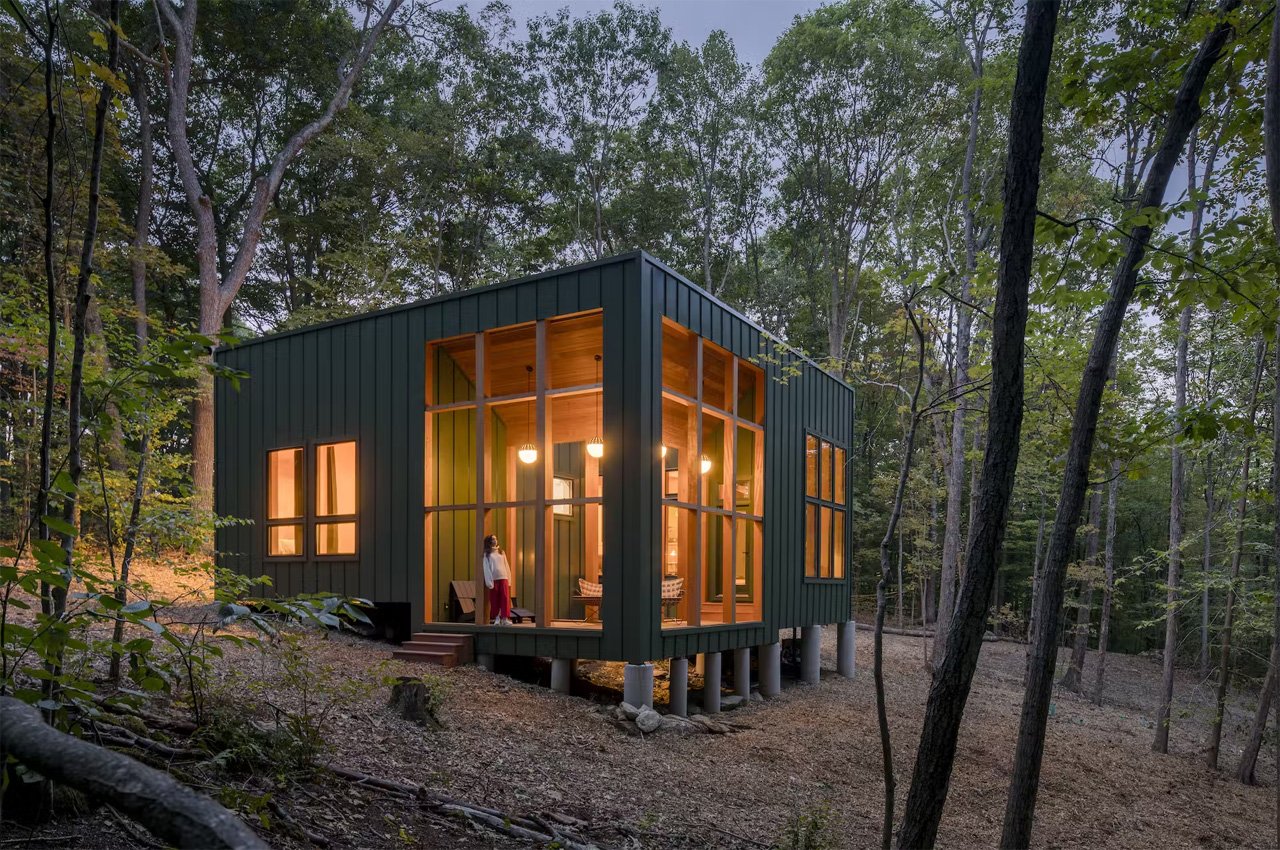 This dark green cabin floats above a sloping terrain in a forest in Connecticut