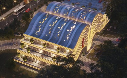 Evening view of a modern building with a wavy blue roof and warm exterior lighting, surrounded by trees and a parking lot.