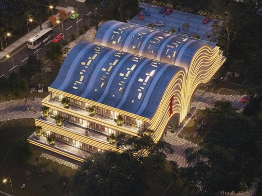 Evening view of a modern building with a wavy blue roof and warm exterior lighting, surrounded by trees and a parking lot.