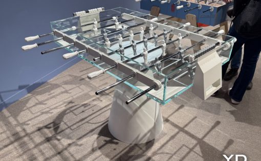 Foosball table with glass sides, metal rods, and white-handled handles in a showroom setting.