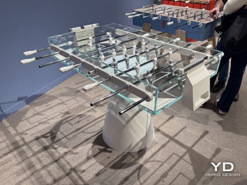 Foosball table with glass sides, metal rods, and white-handled handles in a showroom setting.