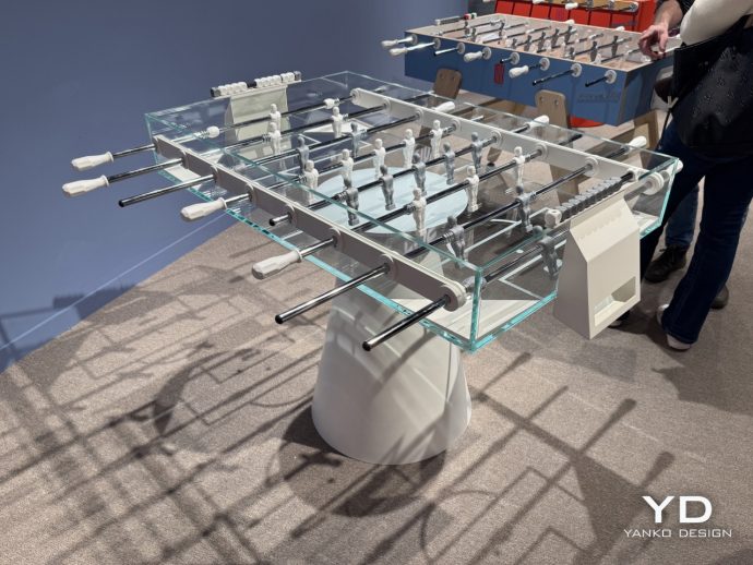 Foosball table with glass sides, metal rods, and white-handled handles in a showroom setting.