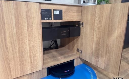 Open lower kitchen cabinet housing a Bosch appliance; a robotic vacuum cleaner sits on a blue mat on the floor in front of the cabinet