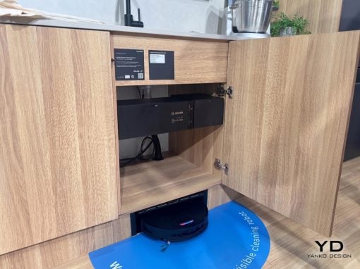 Open lower kitchen cabinet housing a Bosch appliance; a robotic vacuum cleaner sits on a blue mat on the floor in front of the cabinet