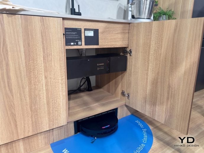 Open lower kitchen cabinet housing a Bosch appliance; a robotic vacuum cleaner sits on a blue mat on the floor in front of the cabinet