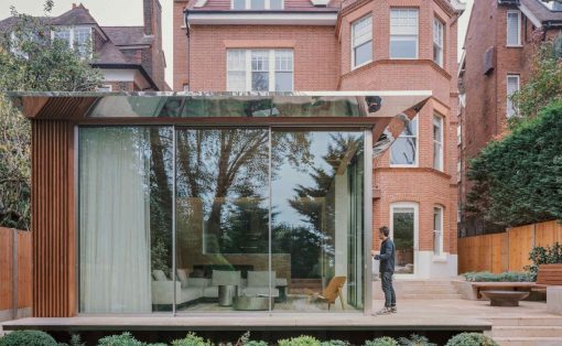 Modern glass extension with wooden slats beside a red brick house; a man stands on the wooden deck near the glass doors.