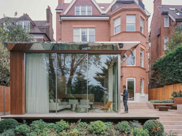 Modern glass extension with wooden slats beside a red brick house; a man stands on the wooden deck near the glass doors.