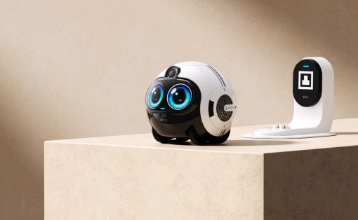 White, spherical robot with big blue eyes sits on a beige pedestal next to a white charging dock/dock station, in a neutral setup.