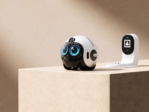 White, spherical robot with big blue eyes sits on a beige pedestal next to a white charging dock/dock station, in a neutral setup.