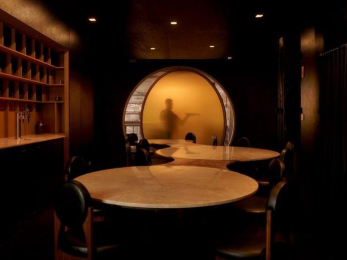 Dim restaurant interior with round marble tables and a glowing circular doorway showing a blurred staff figure in a yellow glow.