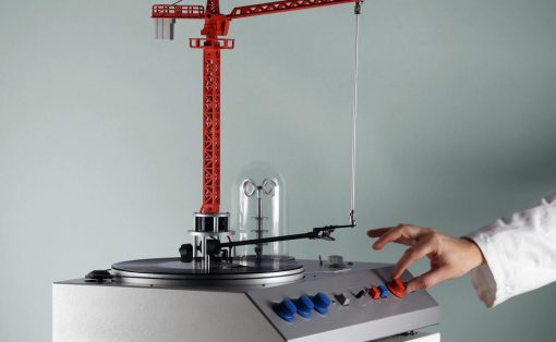 Red construction-crane model mounted on a silver rotary platform, with a hand turning a red control knob on the panel.