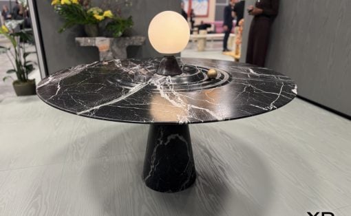 Black marble round table with a glowing orb lamp centerpiece in a showroom; blurred figures in the background and a Yanko Design logo bottom right.