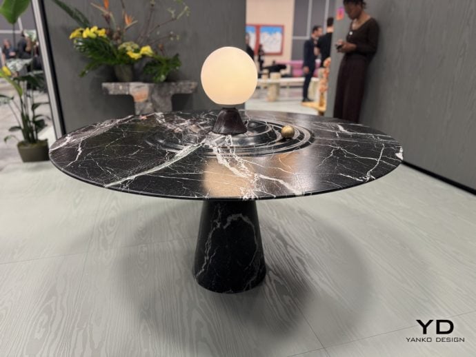 Black marble round table with a glowing orb lamp centerpiece in a showroom; blurred figures in the background and a Yanko Design logo bottom right.