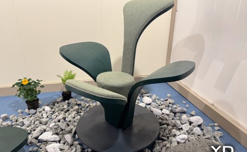 Modern sculptural lounge chair with curved green fabric, mounted on a black pedestal among gray decorative rocks in a showroom setting.