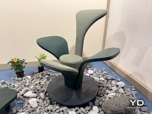 Modern sculptural lounge chair with curved green fabric, mounted on a black pedestal among gray decorative rocks in a showroom setting.