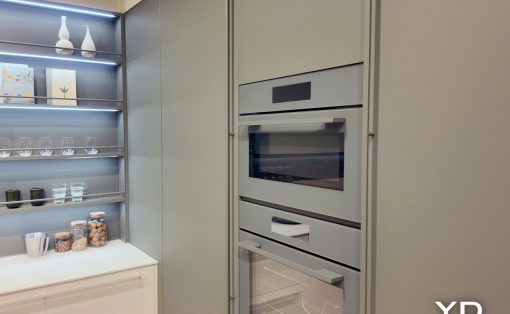 Modern kitchen with a tall built-in oven stack and pale gray cabinetry next to open shelving displaying glassware and jars.