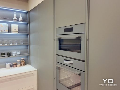 Modern kitchen with a tall built-in oven stack and pale gray cabinetry next to open shelving displaying glassware and jars.