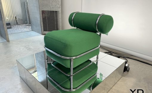 Designer green upholstered chair with a tubular chrome frame on a mirrored pedestal in an industrial studio setting.  The YD Yanko Design logo is visible in the corner.