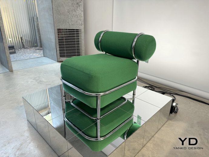 Designer green upholstered chair with a tubular chrome frame on a mirrored pedestal in an industrial studio setting. The YD Yanko Design logo is visible in the corner.