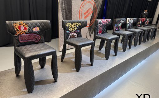 Row of black quilted chairs with colorful embroidered backs on a raised stage at a design event, with a large poster behind.—YD Yanko Design logo visible in the corner