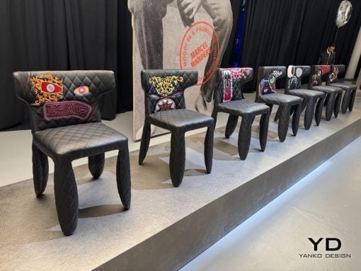 Row of black quilted chairs with colorful embroidered backs on a raised stage at a design event, with a large poster behind.—YD Yanko Design logo visible in the corner