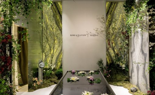Showroom display with a glass water feature containing white water lilies, surrounded by greenery and a branded backdrop.