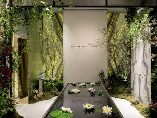Showroom display with a glass water feature containing white water lilies, surrounded by greenery and a branded backdrop.