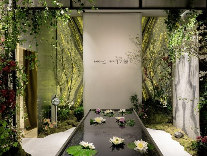 Showroom display with a glass water feature containing white water lilies, surrounded by greenery and a branded backdrop.