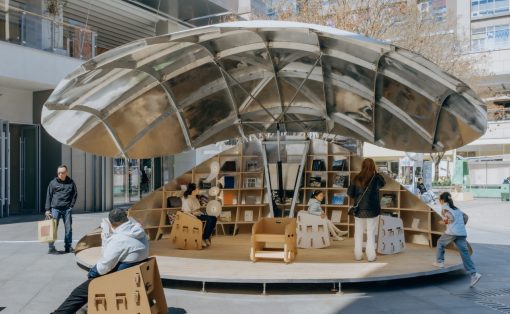 Public reading nook with a large metallic curved canopy and built‑in bookshelves, people sitting and browsing books outdoors.