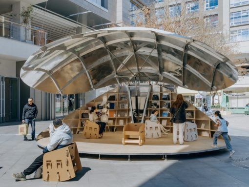 Public reading nook with a large metallic curved canopy and built‑in bookshelves, people sitting and browsing books outdoors.