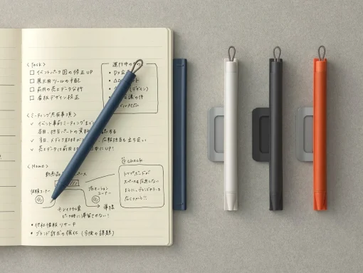 Open notebook with handwritten sketches and checklists, a blue pen resting on the pages; four marker pens (white, black, gray, orange) clipped on a gray surface to the right.