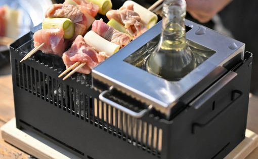 Skewers of meat and green onions grilling on a small portable grill with a bottle placed in a metal holder nearby.