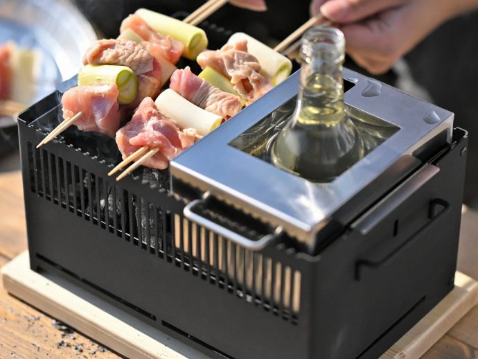 Skewers of meat and green onions grilling on a small portable grill with a bottle placed in a metal holder nearby.