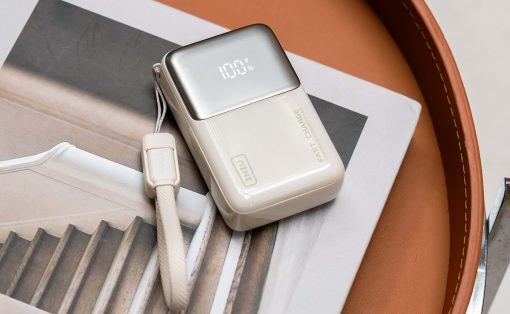 White portable charger with a metallic top showing 100% on its display, placed on a magazine with a white strap nearby.