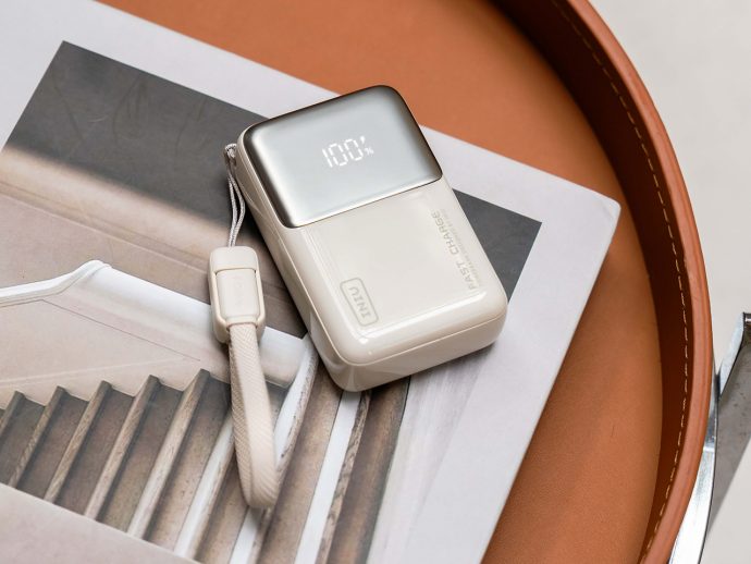 White portable charger with a metallic top showing 100% on its display, placed on a magazine with a white strap nearby.