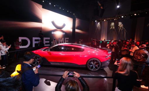 Red sports car on a platform at an auto reveal event, surrounded by photographers and a crowd, with a large DreamME backdrop and screen behind it.