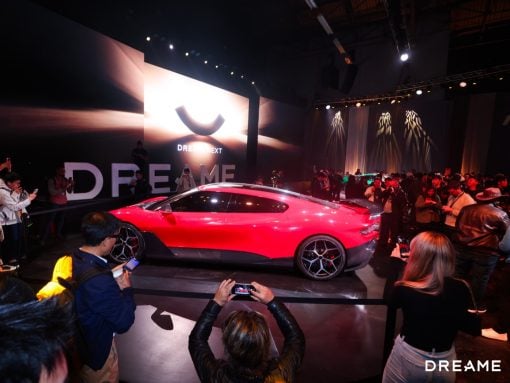 Red sports car on a platform at an auto reveal event, surrounded by photographers and a crowd, with a large DreamME backdrop and screen behind it.