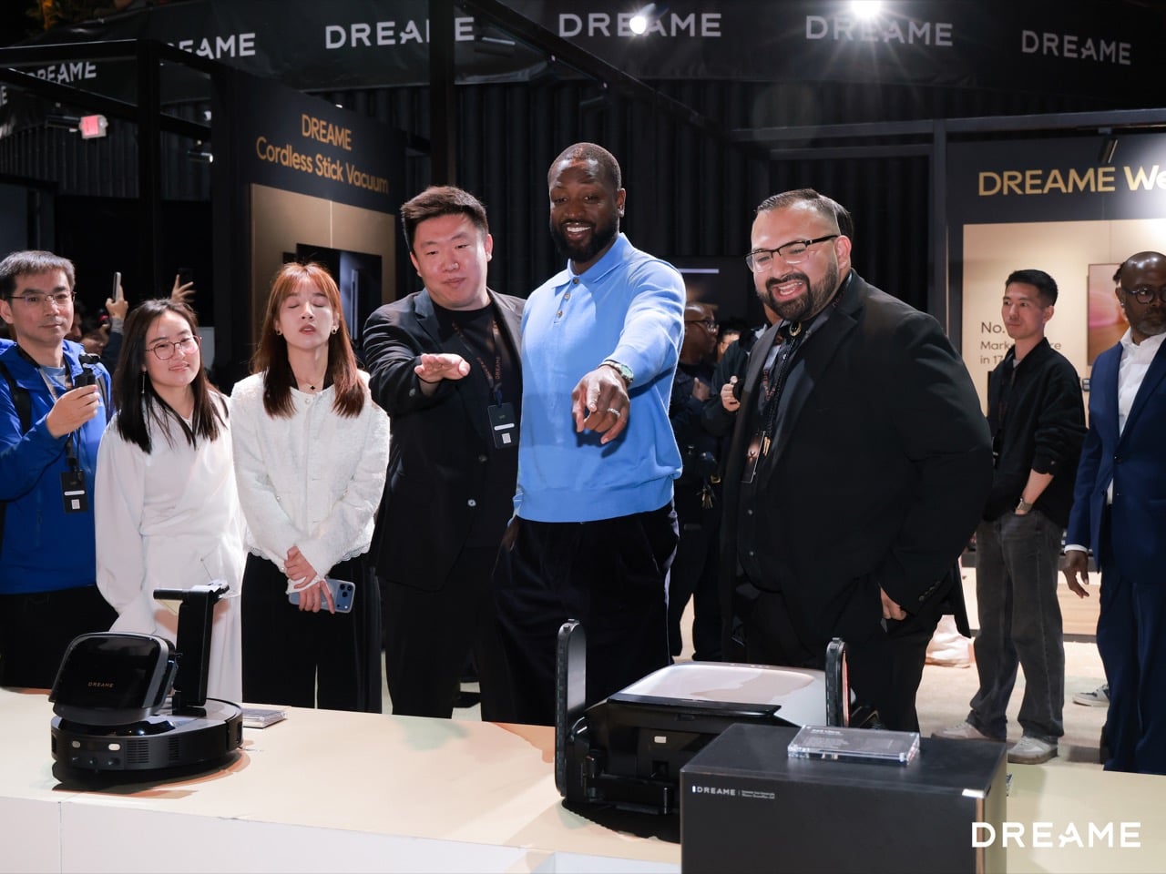 Group of diverse attendees at a DreamE booth, a man in a blue shirt points toward the camera while others smile nearby; a robotic vacuum sits on the table in front of them.