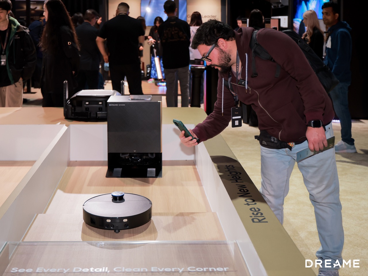 Man in a maroon hoodie leans over a display table to inspect a round robot vacuum while holding a smartphone at a tech expo booth.