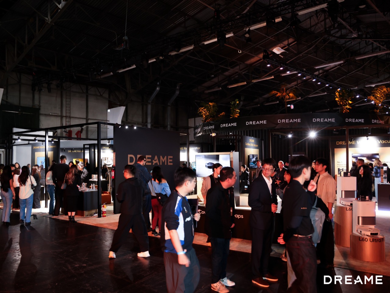 Crowded technology expo with multiple vendor booths and the word DREAME on signage, people chatting and exploring displays at a dark, industrial venue.