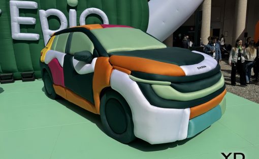 Large inflatable multi-colored car sculpture on a green platform at an outdoor event, with people watching in the background.