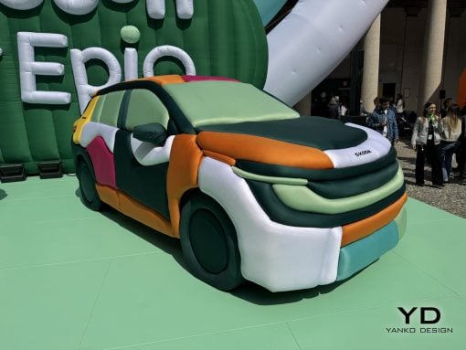 Large inflatable multi-colored car sculpture on a green platform at an outdoor event, with people watching in the background.
