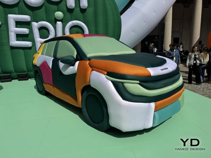 Large inflatable multi-colored car sculpture on a green platform at an outdoor event, with people watching in the background.