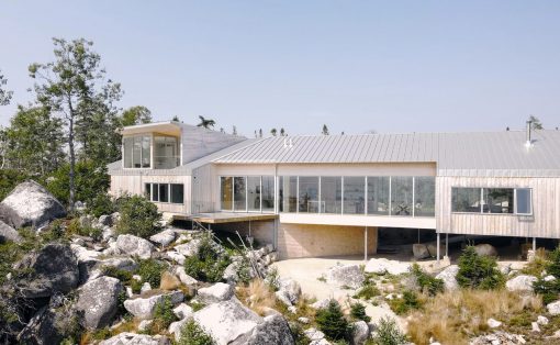 Modern house on a rocky hillside with glass walls and a metal roof, surrounded by boulders and trees.