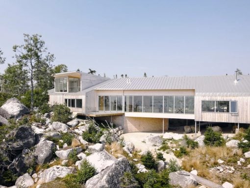 Modern house on a rocky hillside with glass walls and a metal roof, surrounded by boulders and trees.