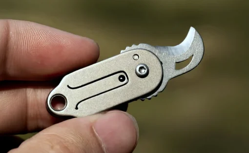 Compact stainless steel keychain multitool held between fingers, showing a curved bottle opener end against a blurred outdoor background.