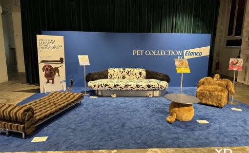 Exhibit booth with a blue backdrop reading 'PET COLLECTION by Elanco'; a dalmatian‑print sofa center stage, flanked by a dark fur chair and a round-topped table on blue carpet.