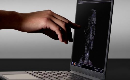 Close-up of a hand reaching toward a laptop screen displaying a fingerprint-shaped pattern made of code characters.