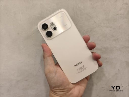 White Honor smartphone held in a hand, showing the rear camera module at the top-left and the HONOR logo toward the bottom on a light background with a watermark in the corner.